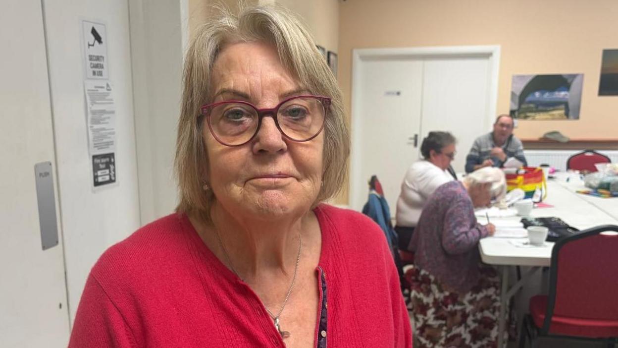 A woman with medium length hair is wearing red framed glasses and a red cardigan. She is standing in a community centre where three people are sat at a white table in the background.