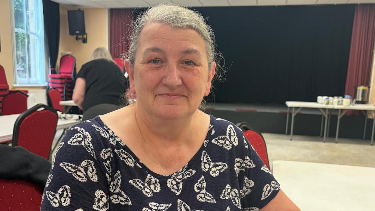 Alison Grant had grey hair tied back and is wearing a black t-shirt with white butterflies on it. She is sat next to a table in a community hall. In the background there is a stage and tea and coffee making facilities.