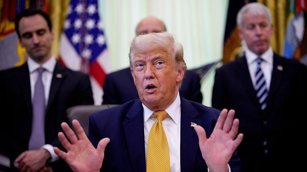 US President Donald Trump wearing a navy suit, white shirt, and gold tie holds both of his hands up as he speaks during an event in the Oval Office of the White House. Two mean in suits and ties are on located on the left and right of him.