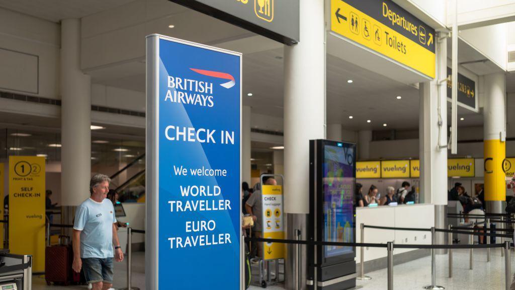 A check in area at London Gatwick has a sign for British Airways. Staff are being a desk and a man in a T-shirt is near a sign that says self check-in. There is a screen near the check-in desks. A yellow sign overhead points to Departures.