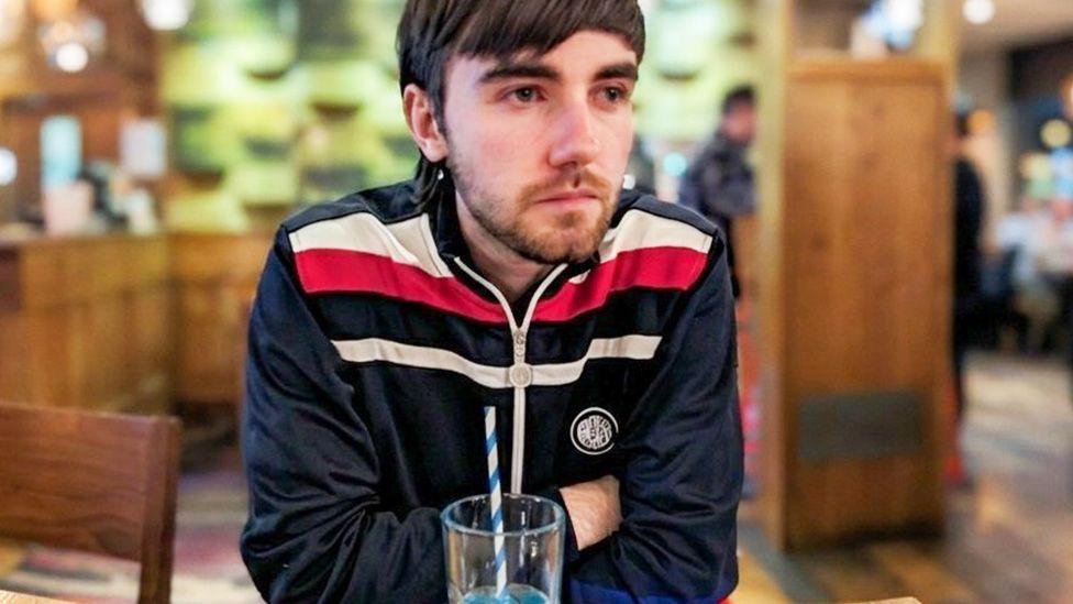 Nathan sitting in a pub with a drink. He is wearing a blue tracksuit top and has his arms crossed. He has a short beard and dark brown hair.