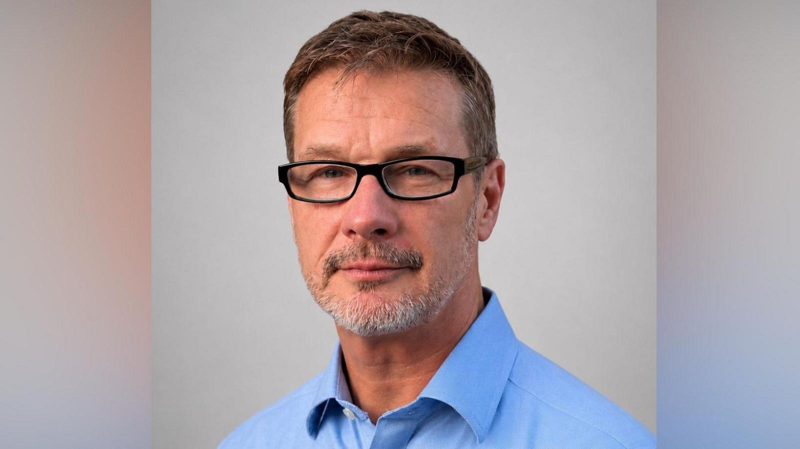A man in a blue shift with glasses. He has grey stubble.