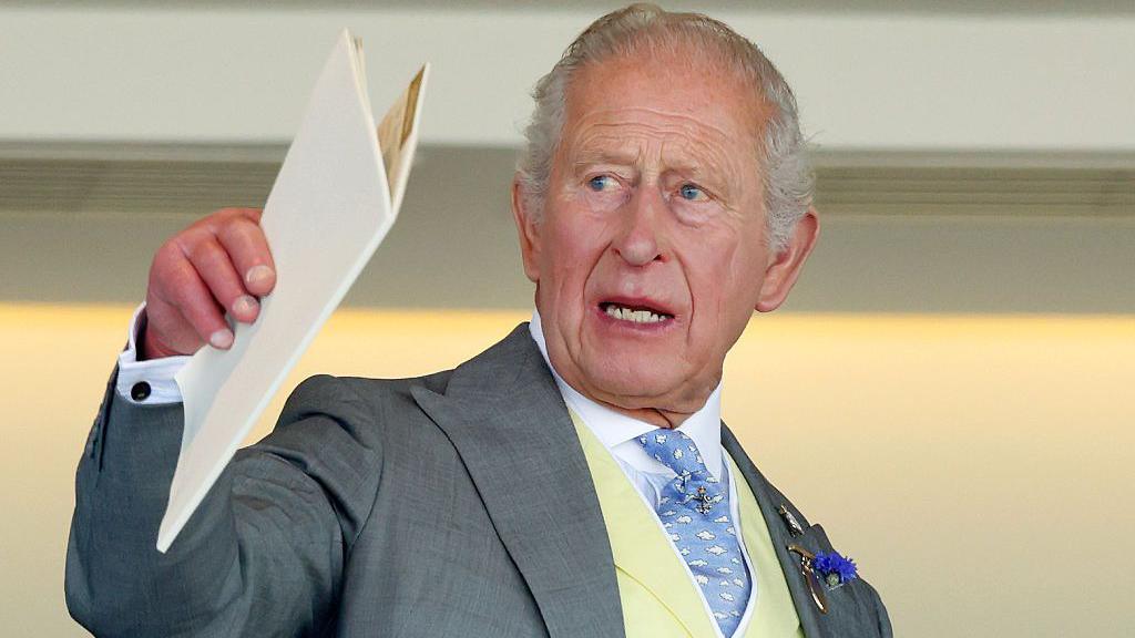King Charles holding a white A4 booklet and gesturing with it. He is wearing a pale great suit, cream waistcoat, blue and white patterned tie, and a white shirt.