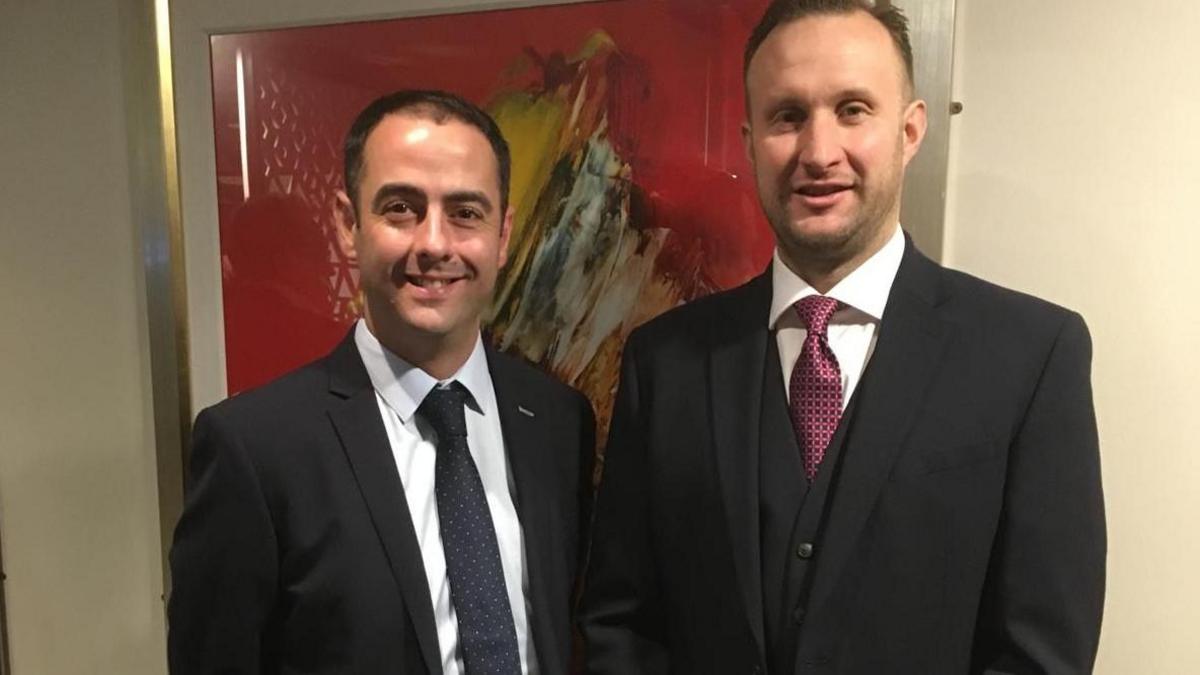 Two men with short brown hair are wearing a shirt and tie and smiling at the camera.