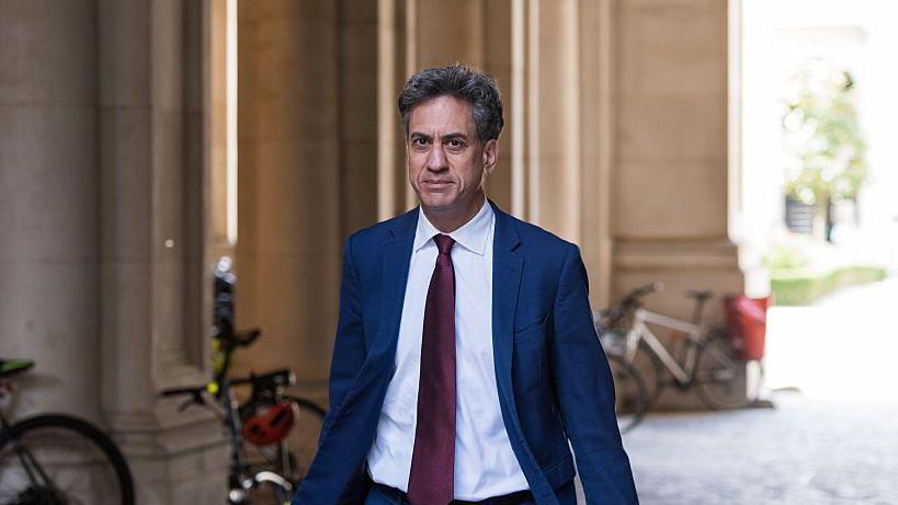 Ed Miliband walking down a cobbled pedestrianised street in London. He is wearing a white shirt, blue suit, and red tie.