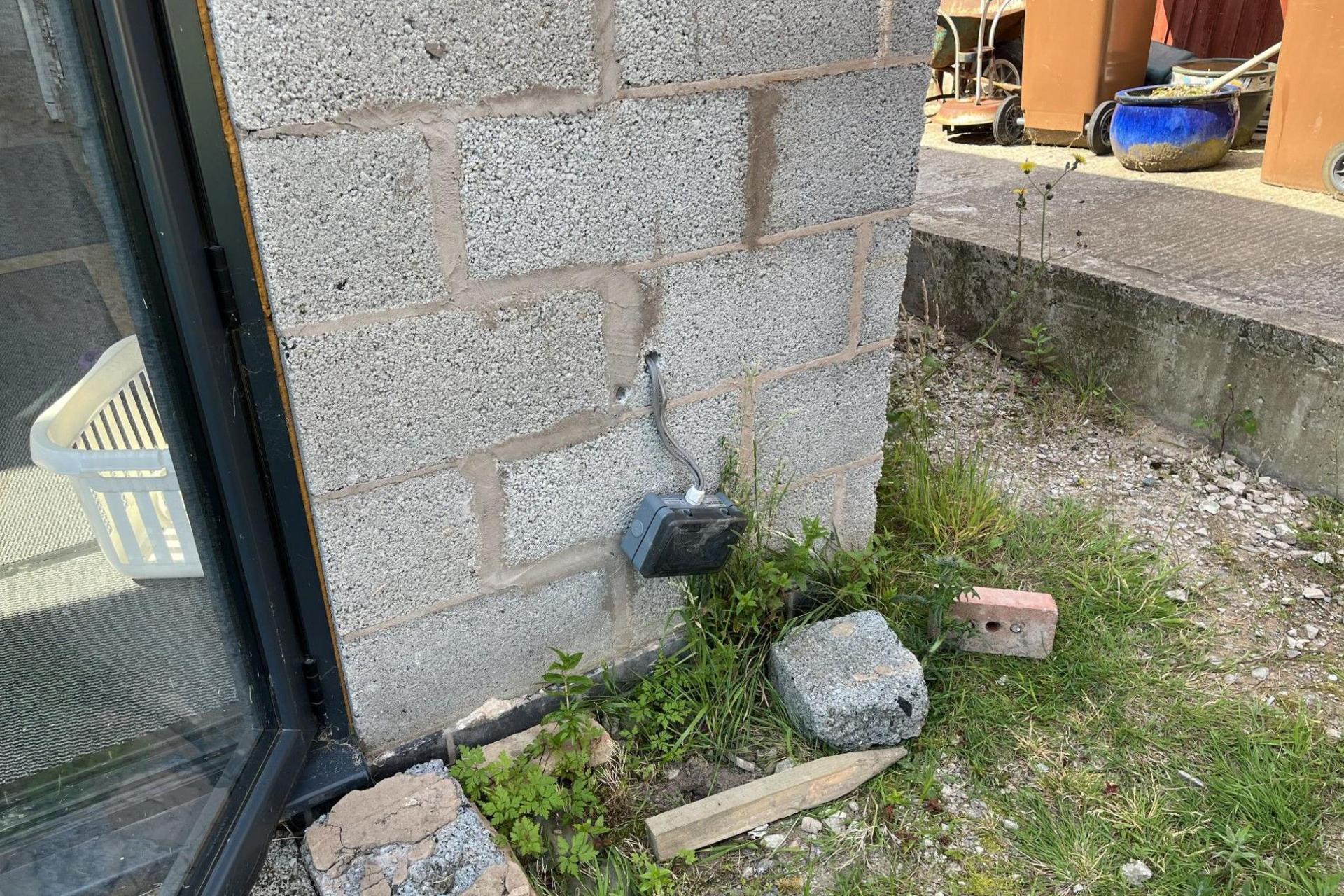 Close up of a corner of a building that has exposed grey breeze block and building rubble on the ground. There is an outside electrical socket dangling off the wall.