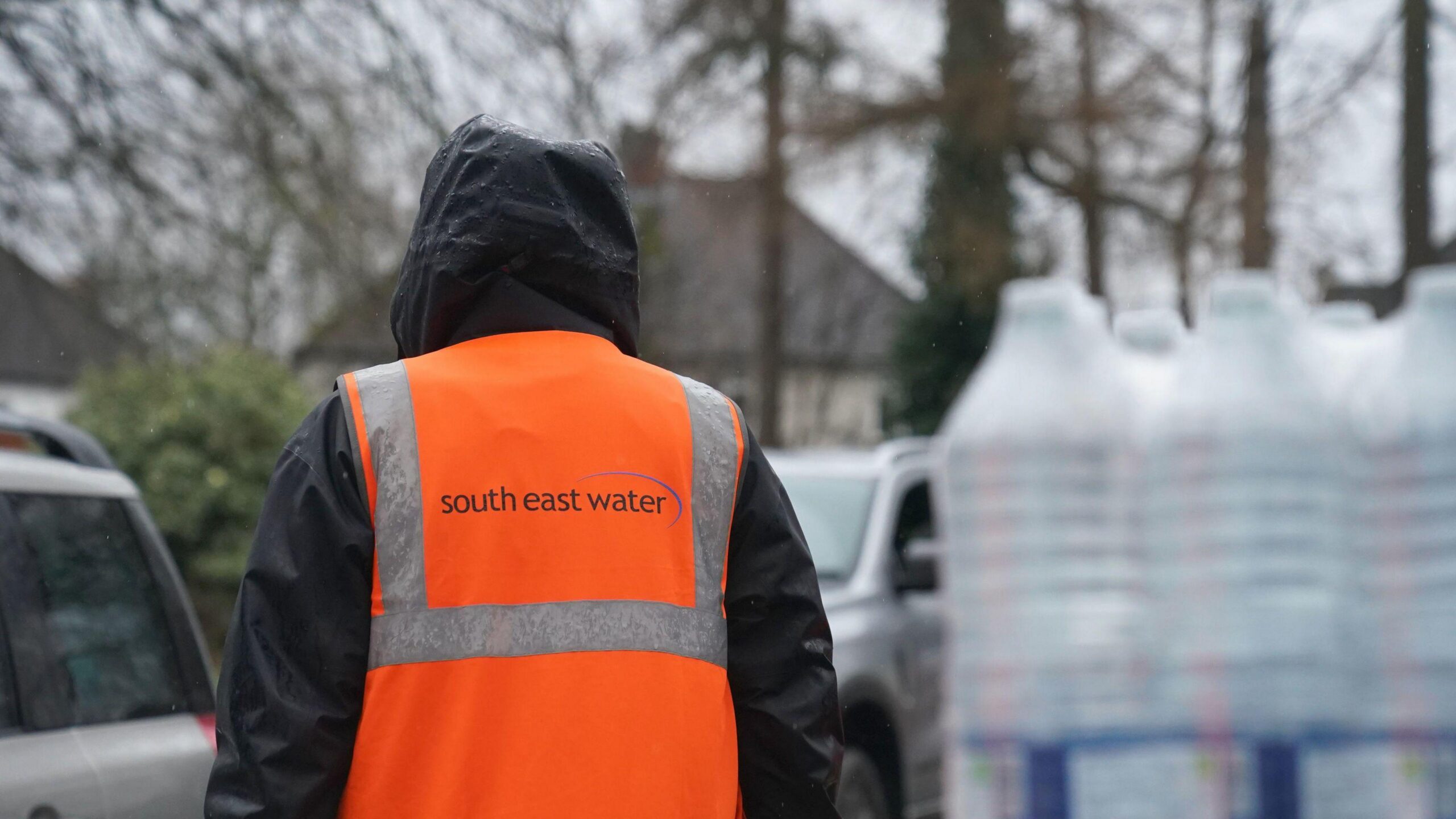 A person stands with their back to some bottles of water. The sky is grey and it is drizzling. They have the hood of their water-proofed jacket up.