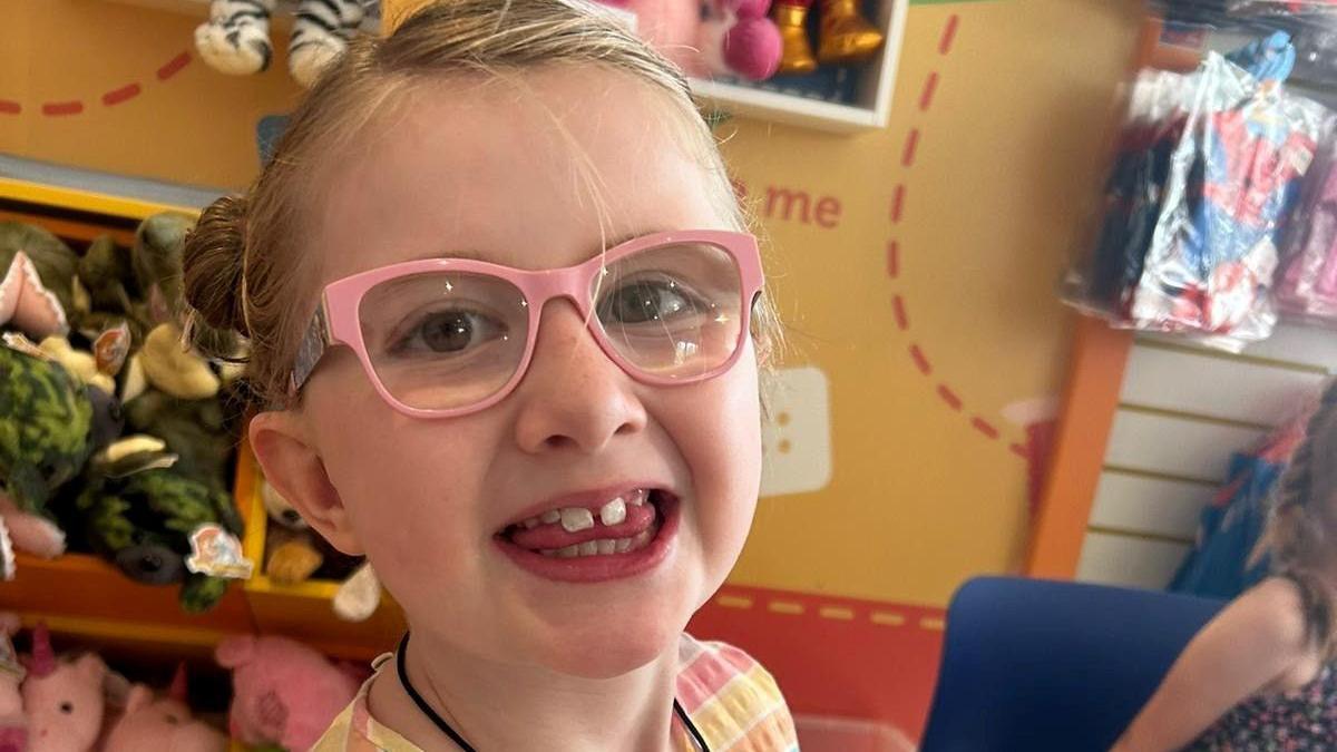 A sweet girl in pink glasses and blonde hair tied back smiles to the camera
