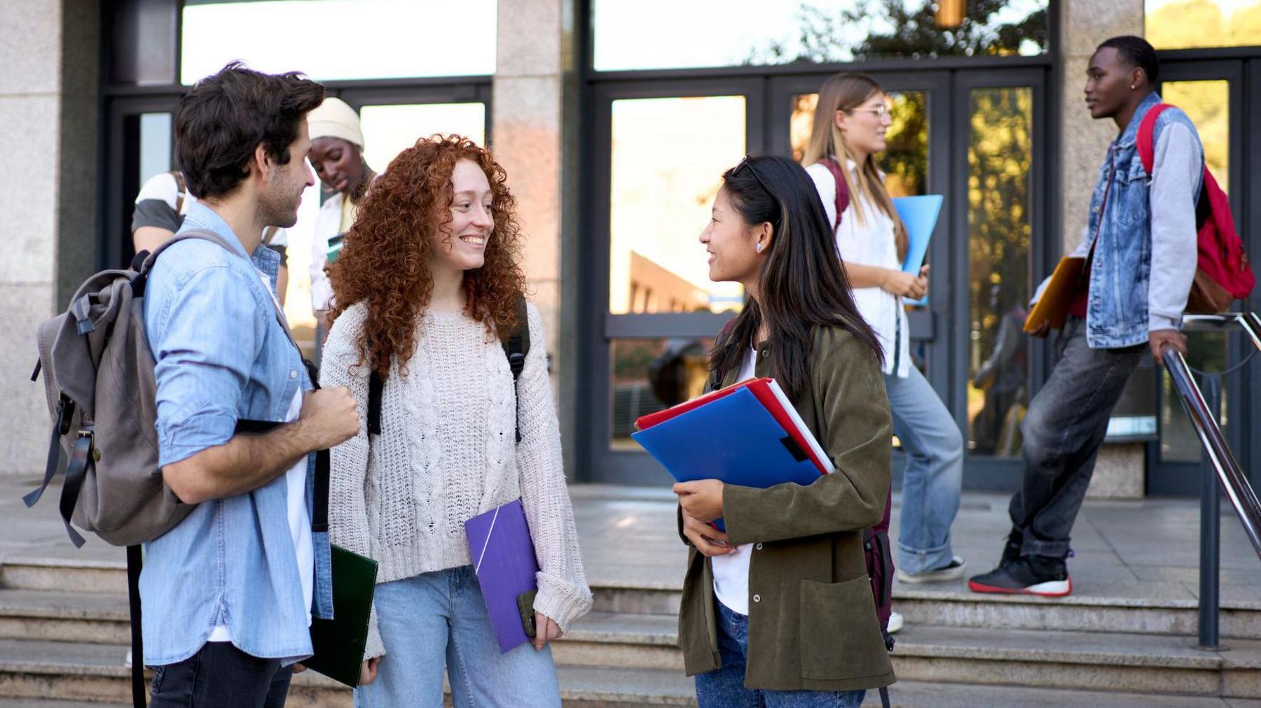 A group of students stand chatting on steps outside a campus building, carrying backpacks, notebooks and folders, while others walk past the entrance behind them.