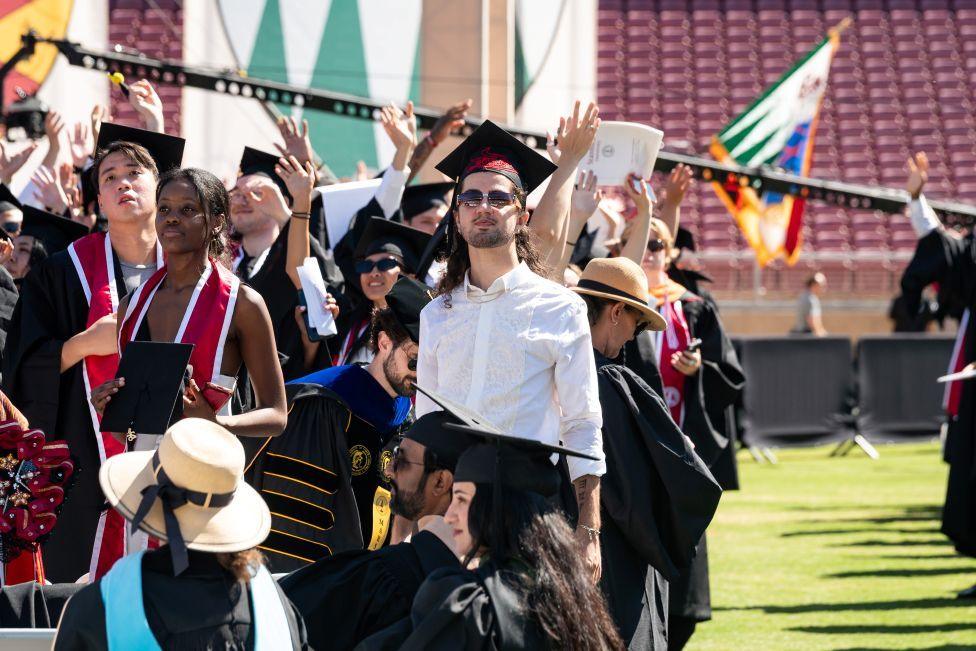 Atash is dressed in a white shirt and black mortar board and sunglasses. He is surrounded by other students some of whom have their arms in the air.