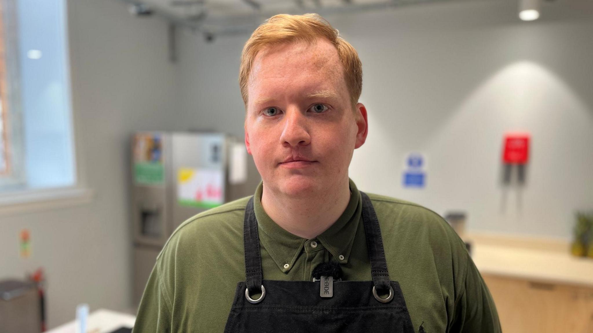 Peter Gilchrist, with ginger hair and wearing a green T-shirt and black apron, is standing in an industrial kitchen.
