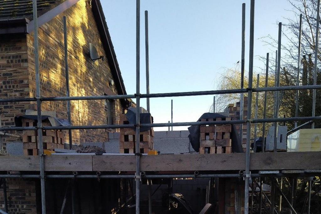 An image of a house with a half built extension on its right hand side. The extension is one storey tall with scaffolding all over and piles of bricks on top ready for the next level.