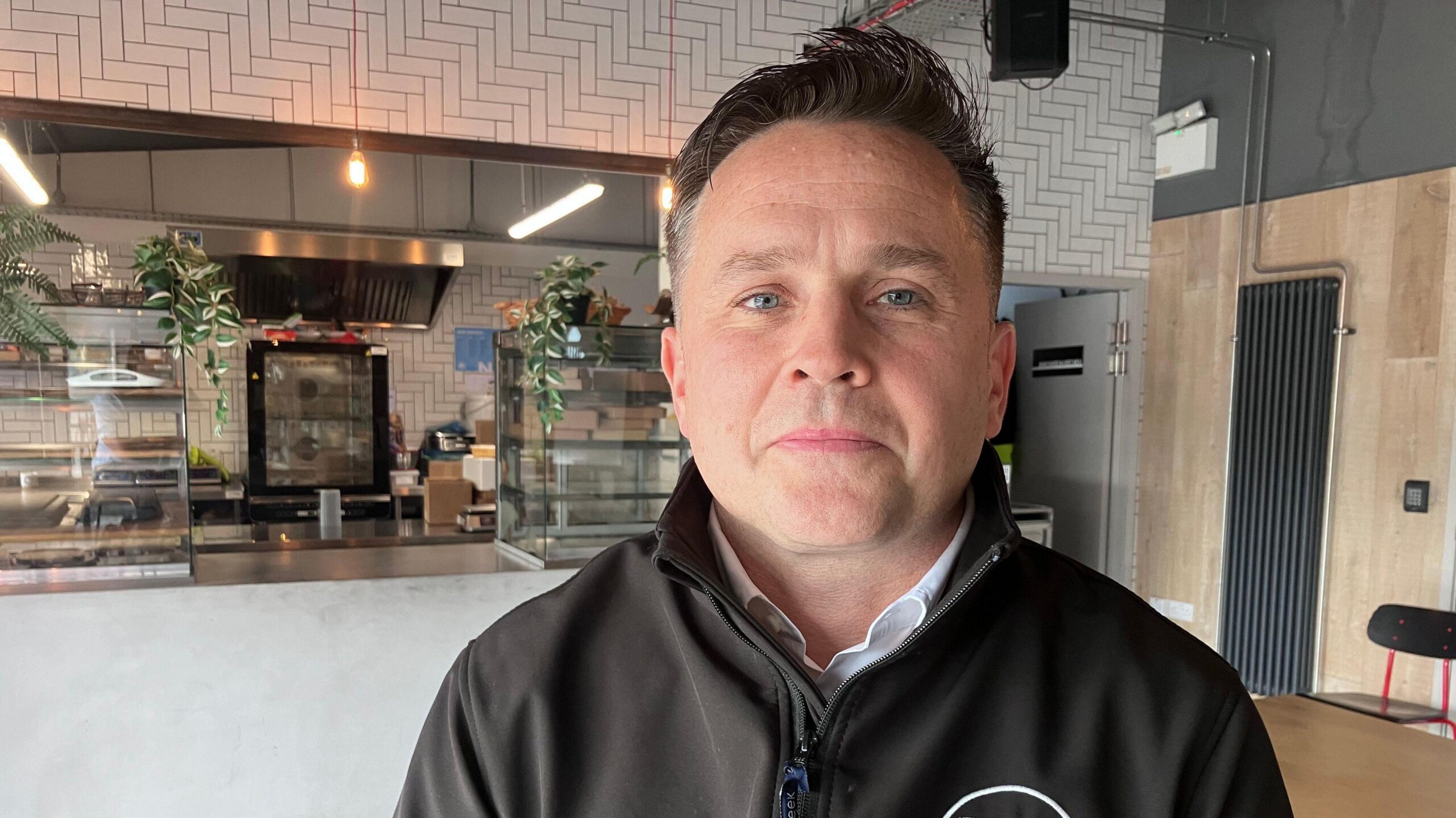 Brian Reid. He has dark hair and blue eyes, wearing a black zip up jacket with white shirt underneath. A cafe counter is behind him.