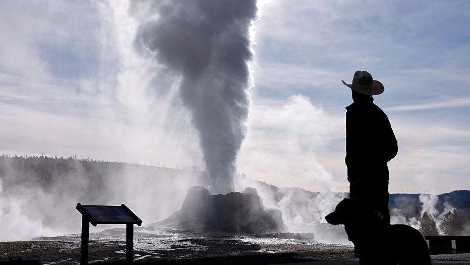 A man is a cowboy hat and his dog are silhouetted in front of the steam coming from the Castle Geyser in Yellowstone National Park, Wyoming.