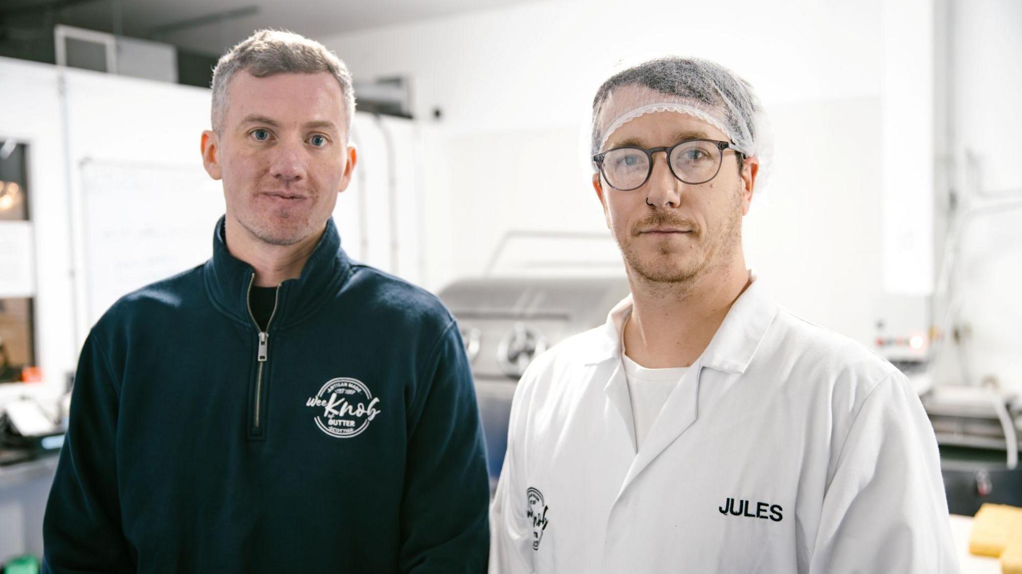 Kieran Woods is wearing a blue fleece with the Knob of Butter logo on it. He is standing next to Jules Bal who is wearing a hair net, glasses and a white T-shirt with a white overall over it. It has the name Jules sewn into it.
