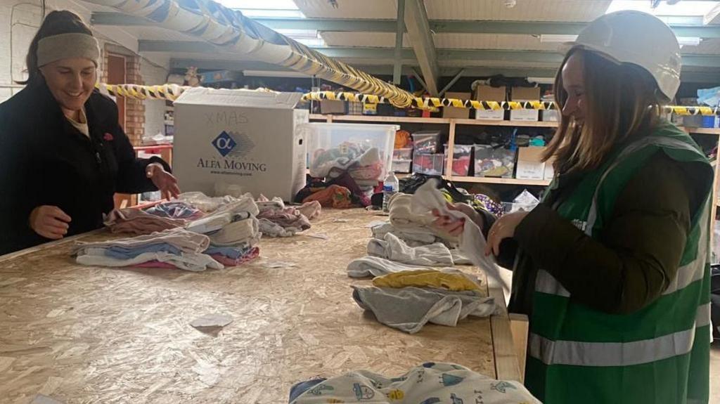 Two women inside a large industrial unit, sorting baby clothes out. The woman on the right is wearing a hard-hat with a green high vis jacket and the woman on the left is hearing a head band, and a dark top. They are both smiling. There are baby items on a wooden desk and rows of boxes on shelves behind.