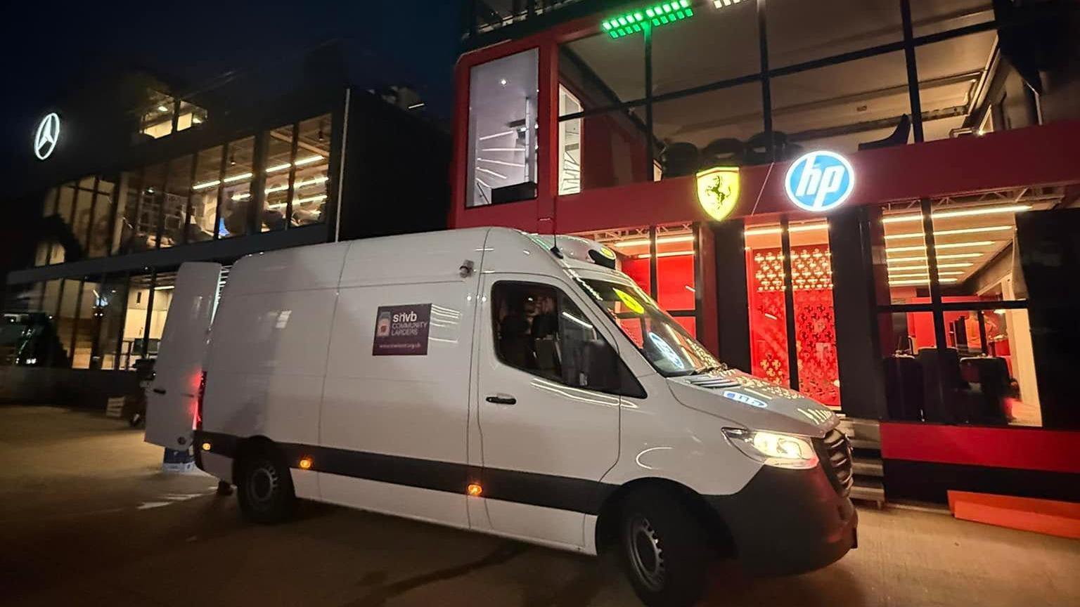 A white van is parked at Silverstone outside two buildings, one displaying a lighted Mercedes sign and the other a lighted Ferrari sign.