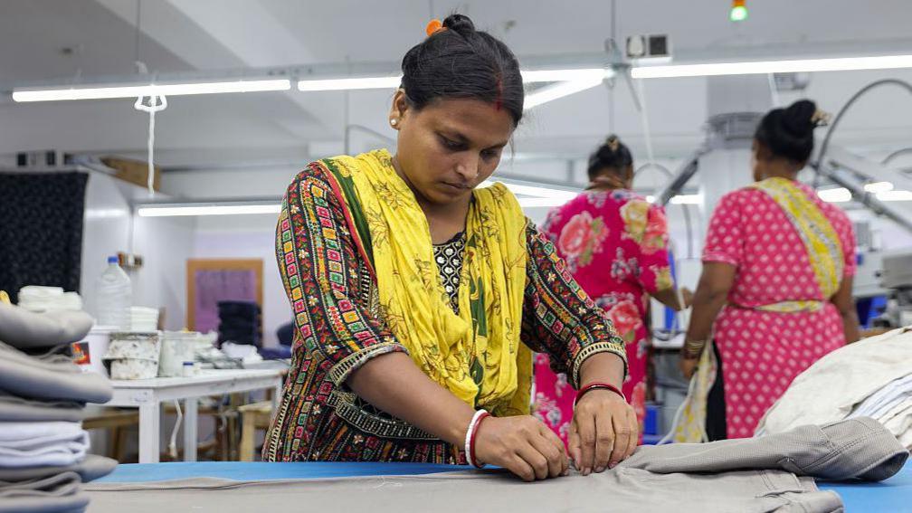 A woman wearing a multi-coloured Indian kurta with a yellow stole, finishes men's trousers after production in Kolkata city, India,.