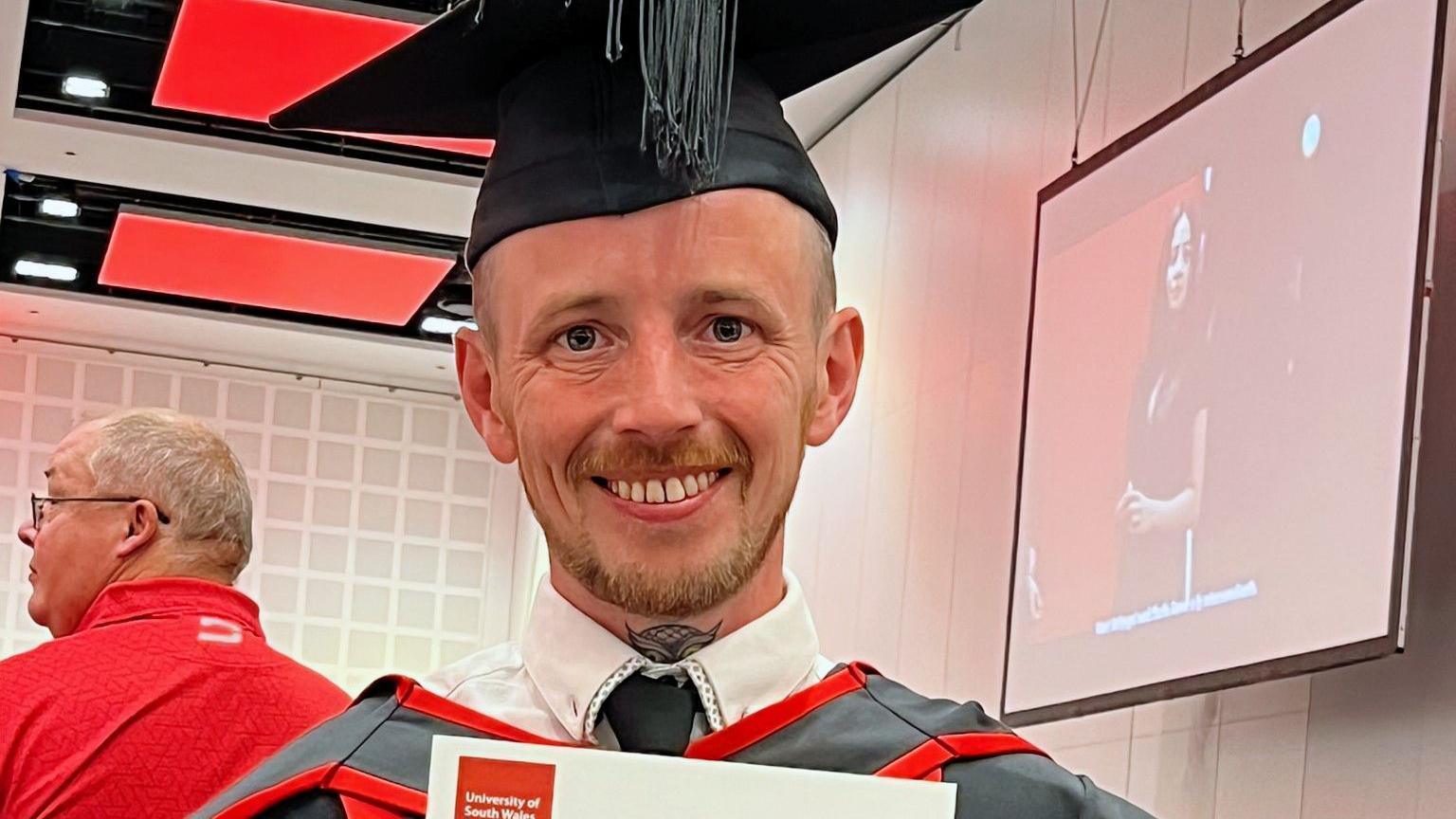 Steven, a man in a mortar board hat and academic robes holding up his graduation certificate. He has a short ginger goatee.