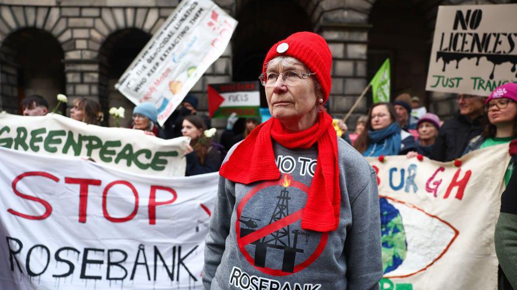 Protesters outside Edinburgh's Court of Session with white banners saying "stop Rosebank". In the foreground is a woman with glasses, a red hat and scarf, and a grey jumper with an anti-Rosebank slogan