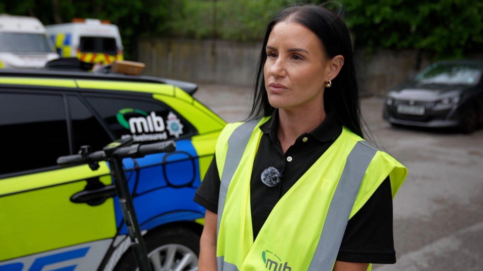 Hayley Sutcliffe from the Motor Insurers' Bureau is standing in front of a police car and an e-scooter, she has dark shoulder length hair and is wearing a high-vis vest.
