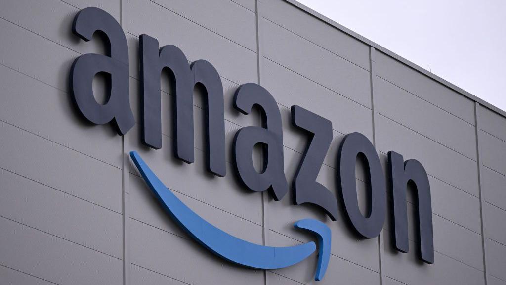 The Amazon logo, which is the world 'Amazon' with its signature stylised arrow underneath, on the side of a grey building with a light overcast sky in the background