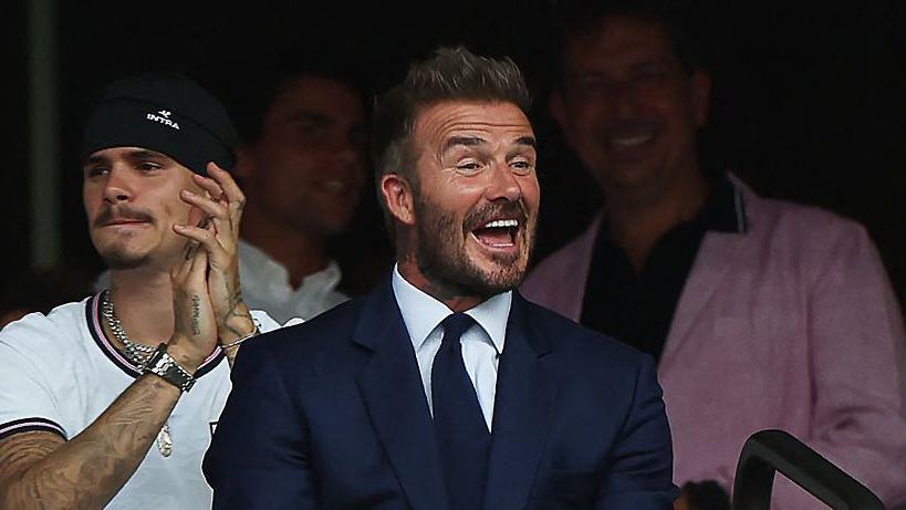 English former football player David Beckham smiles during the 2026 World Cup football tournament quarter-final match between Norway and England at Miami Stadium in Miami Gardens on July 11. He is wearing a blue suit, white shirt and a blue tie.