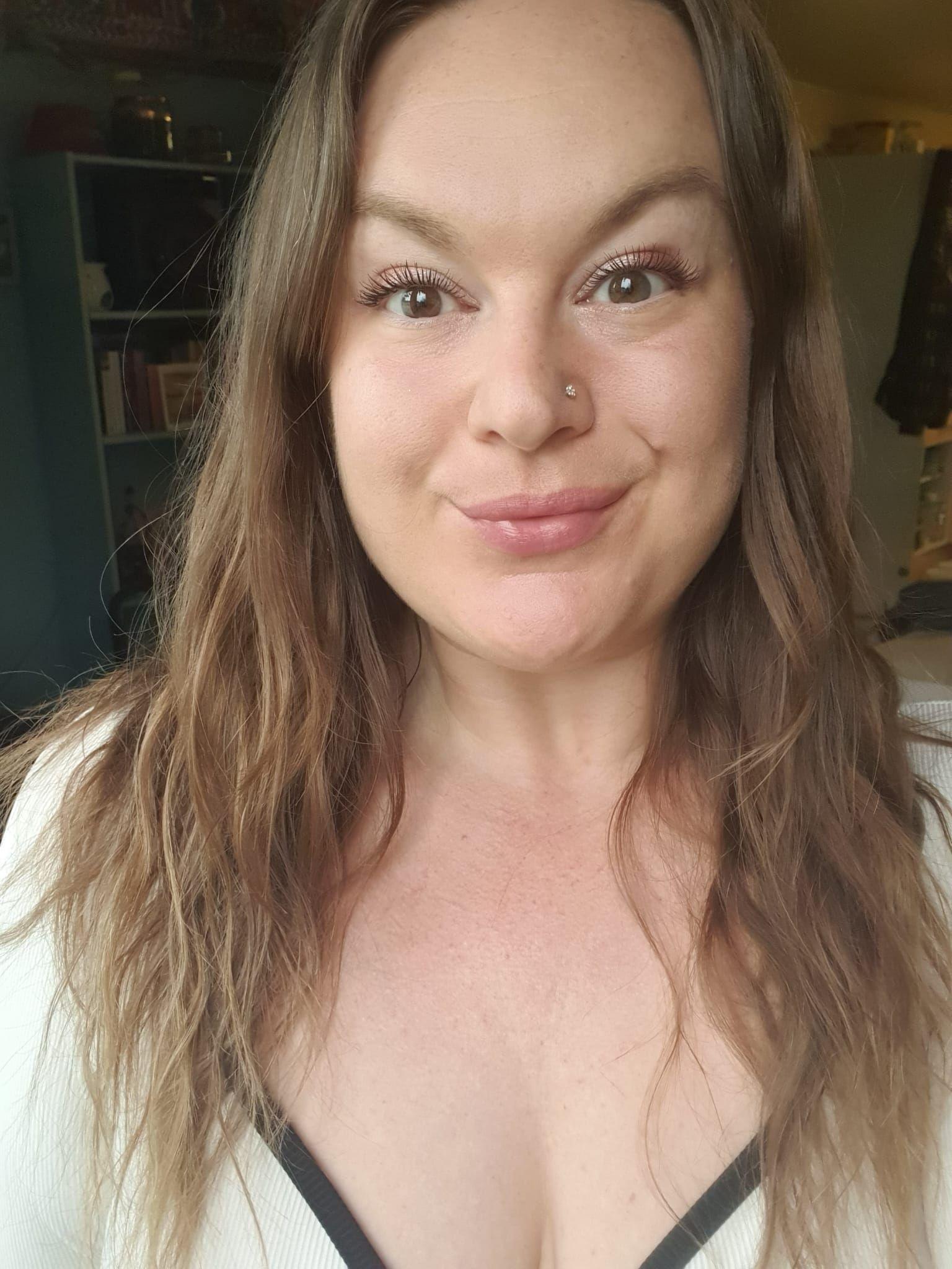 Selfie of a young woman with long brown hair, brown eyes and a nose piercing smiling. She wears a white top with black trim.