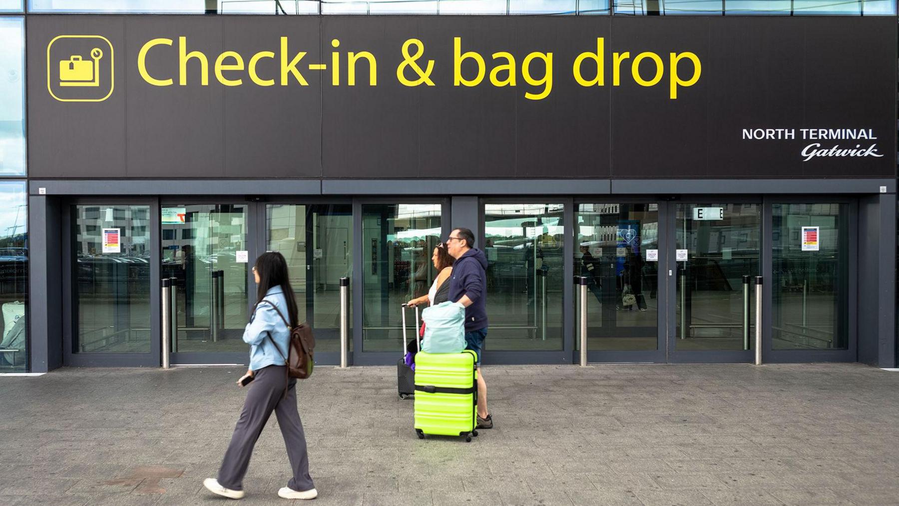 A sign outside Gatwick airport reads 'check-in & bag drop; north terminal Gatwick', while people walk past wheeling baggage along the concrete floor, image taken in 2024.