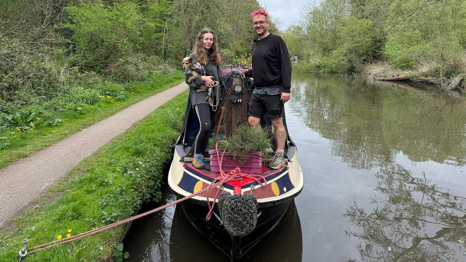 A young man with pink hair and glasses stands on a narrowboat moored on a river next to a young woman with long wavy hair who is holding a small dog. There is a towpath to the left of them.