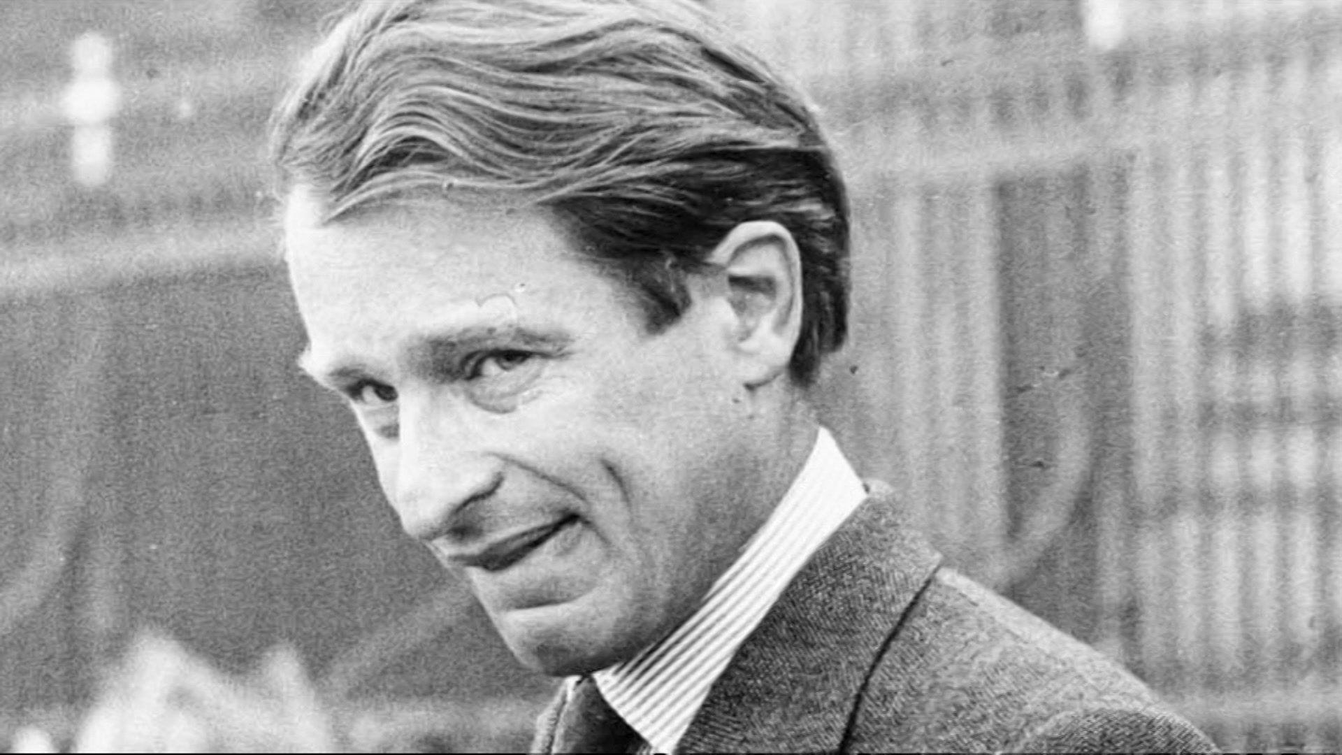 A black and white image of a man with wavy trimmed hair swept back from his forehead. He is looking sideways at the camera and appears ot be dressed smartly.