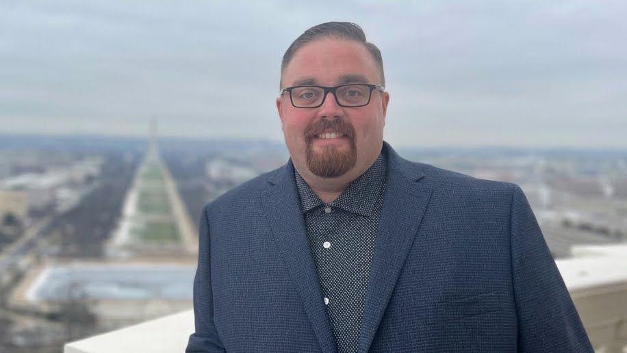 Michael Fewell wears a blue suit and grey shirt with the outline of a city behind hi.
