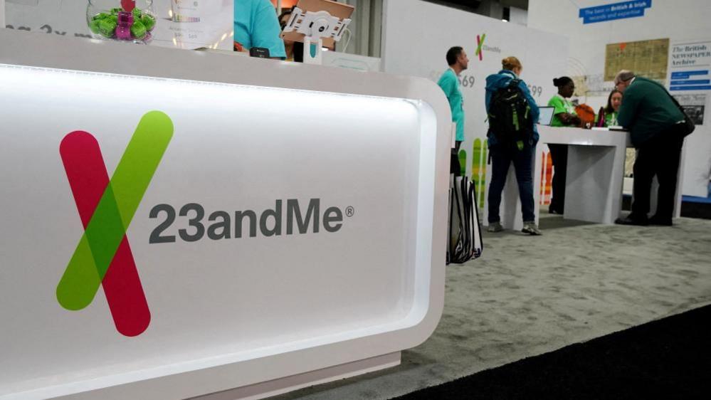 The 23andMe logo is seen at a genealogical convention. The logo is on a bright, white display stand at the front of a carpeted exhibition stall for the company, with people in light blue uniform shirts and others in plain clothes around a table in the background.