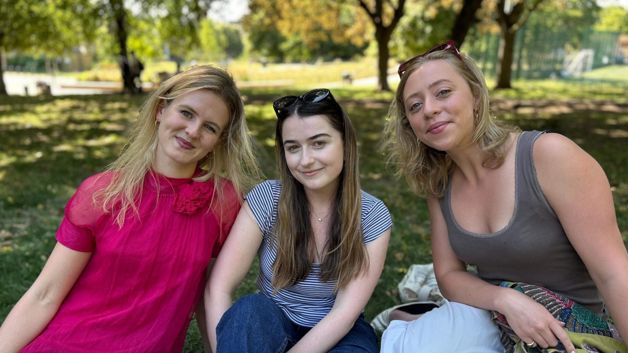 Group of three women sat in park, all smiling at the camera. The woman on left wears a fuchsia pink dress, the woman in the middle, a stripy t-shirt and jeans and then the woman on the right, a sage green vest top. Behind them is a park with leafy trees.