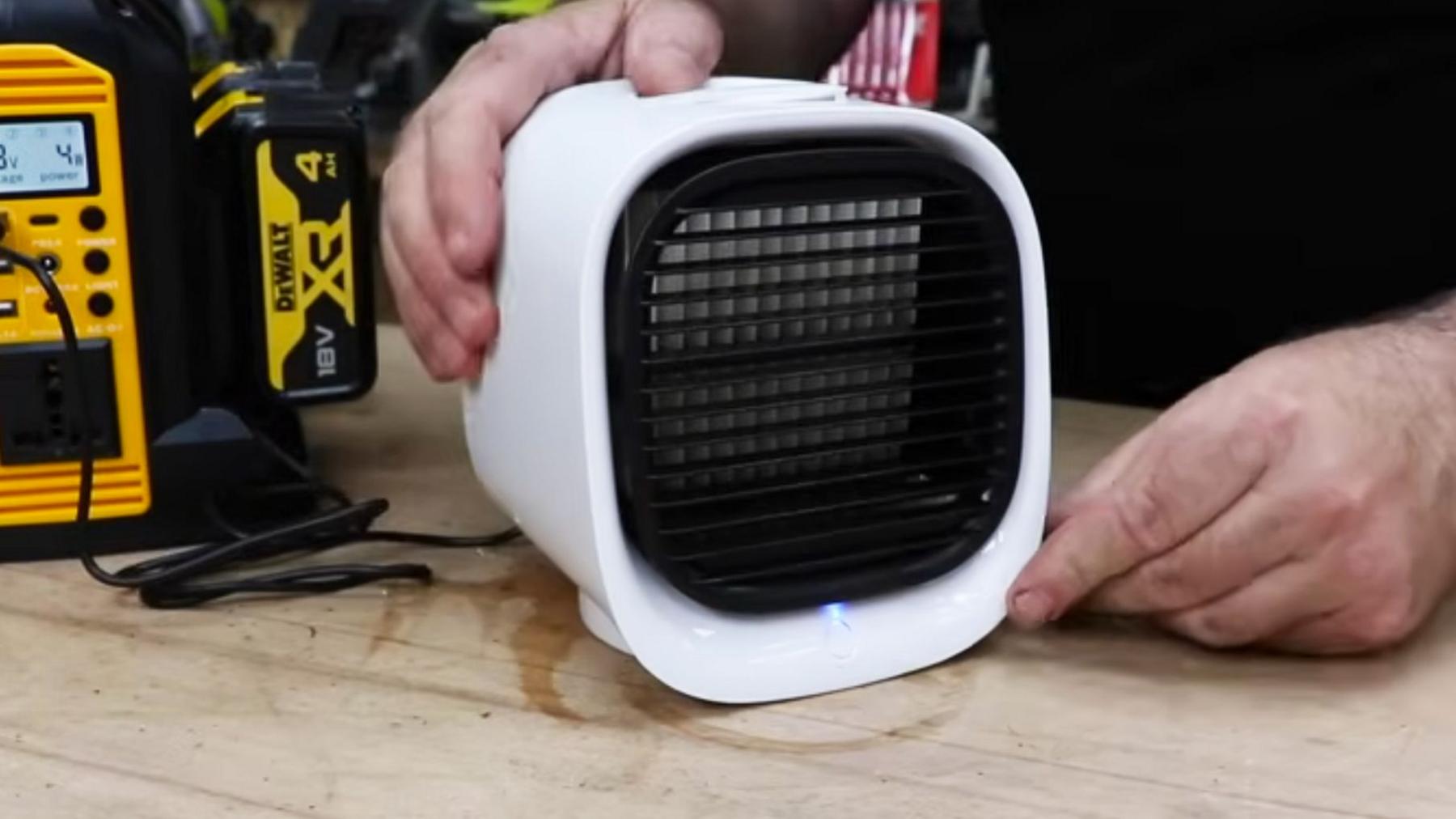 A small cube shaped white portable fan which claims to be an air conditioning unit on a desk with hands holding it