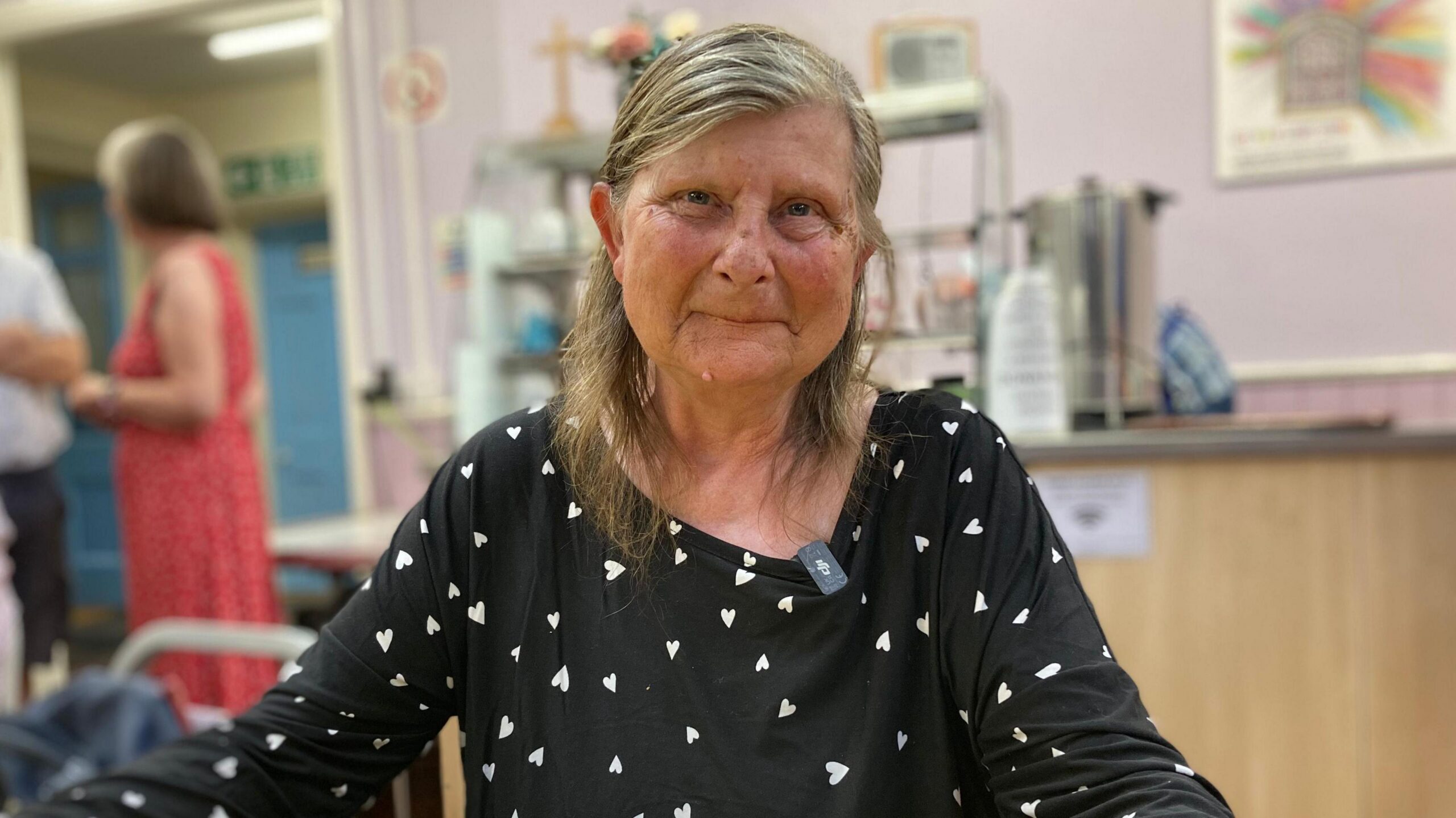 A woman smiles at the camera as she sits in a cafe shop. She has grey shoulder-length hair, tucked back behind her ears, and is wearing a black blouse covered in small white hearts.