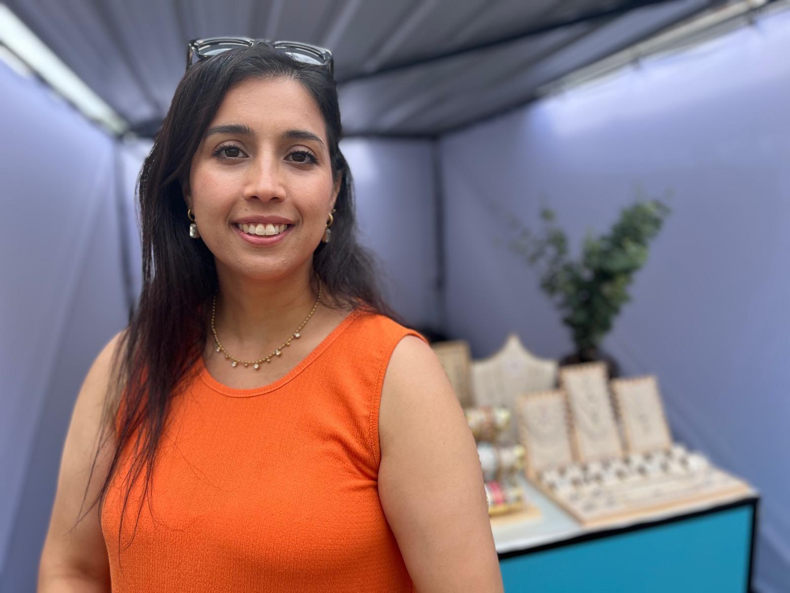 Buket Sevic standing to the left of the shot wearing an orange vest with long dark straight hair and black sunglasses on her head. To the right of the shot behind her is a selection of jewellery