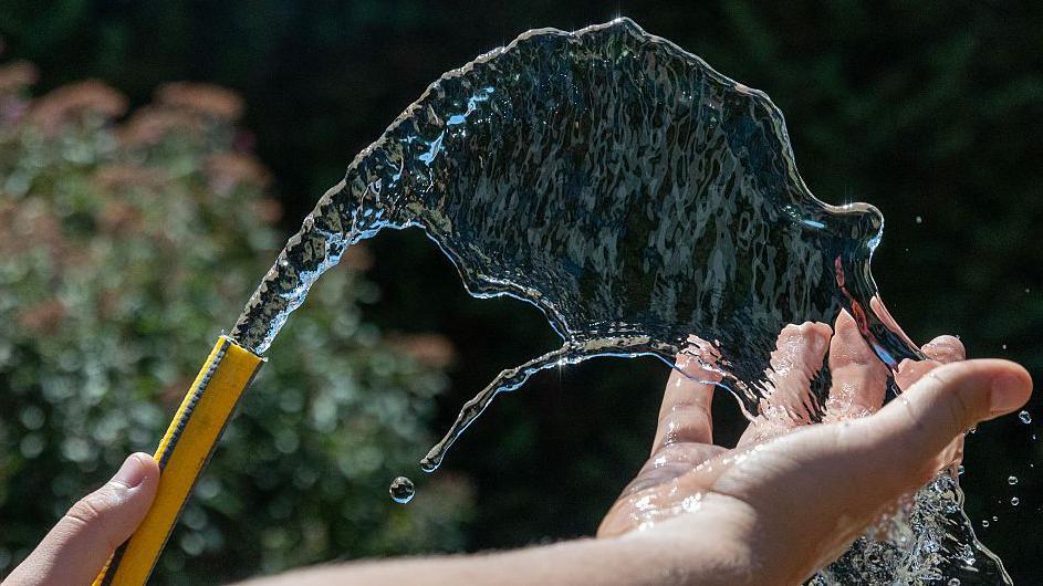 A person using a yellow garden hose which is spraying water into the air and on to someone's hand.
