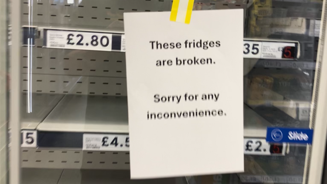 A sign saying 'these fridges are broken, sorry for any inconvenience'. There are empty fridge shelves behind it.