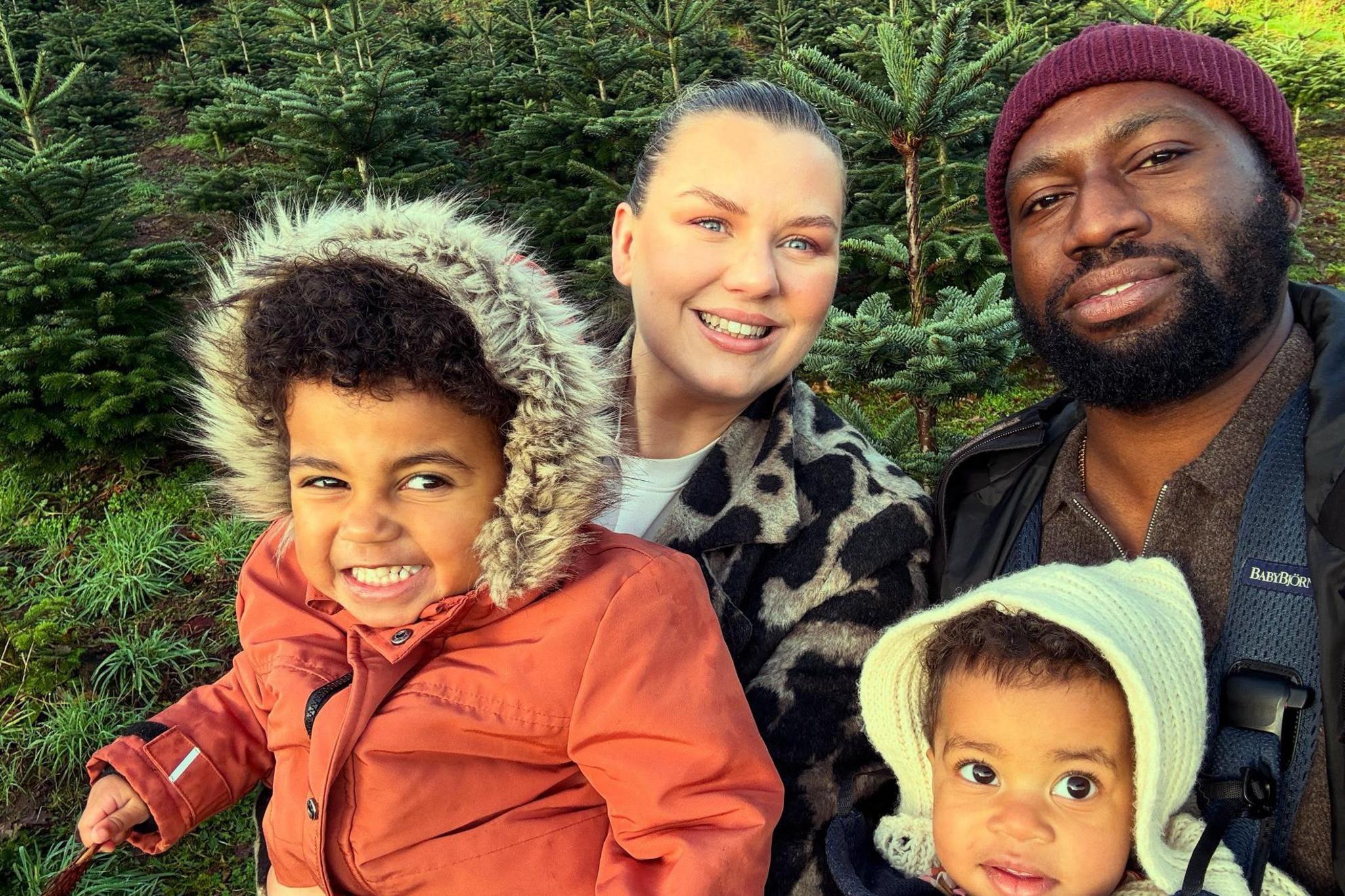 A family of four taking a selfie - the kids are wearing warm winter clothes - and the man and woman are stood behind. Looks like they are in a Christmas tree farm.