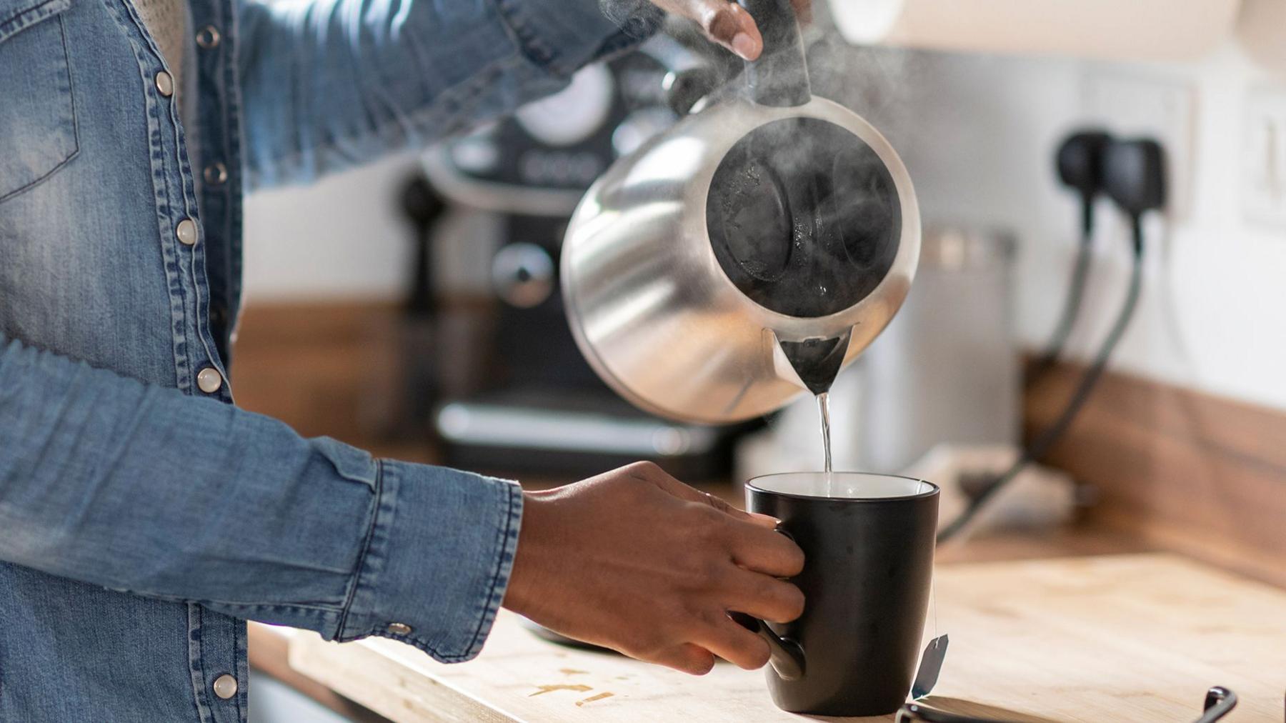 A women pours water from an electric kettle into a mug. A plug socket can be seen in the background