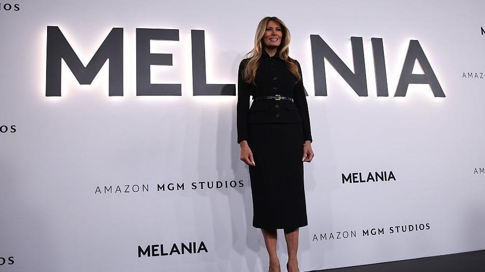 Melania Trump stands in front of the title 'MELANIA' in large letters at the premier of the documentary film