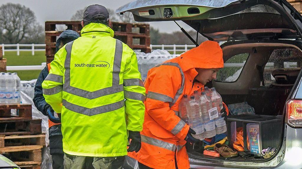 Man in orange hi vis putting bottles of water in boot of car