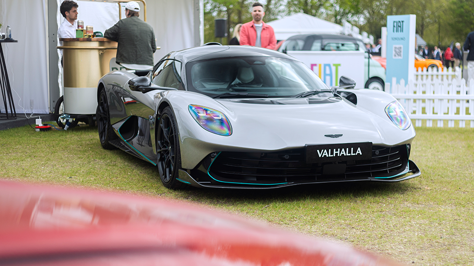 A silver grey Aston Martin Valhalla on grass, at an event. There arethree people in hte background