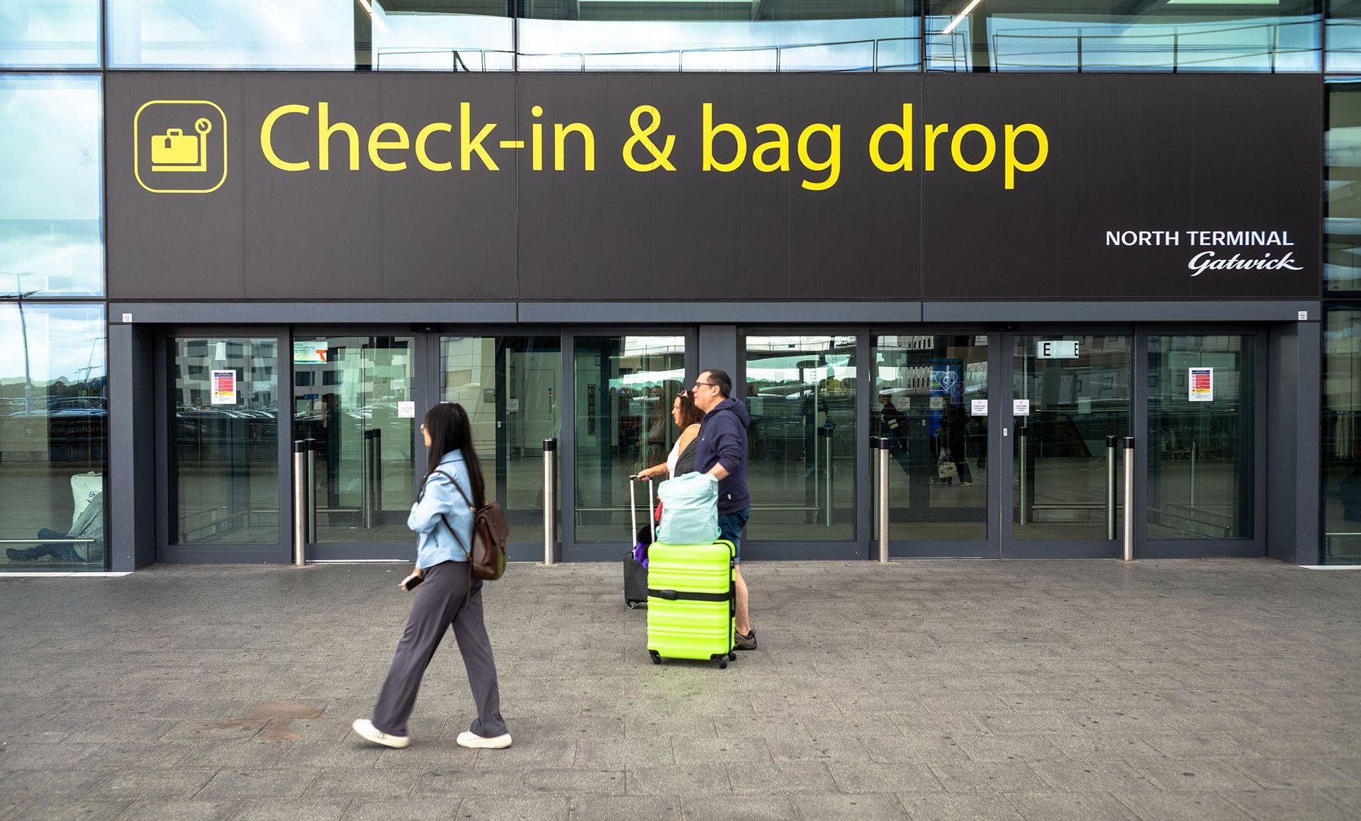 A sign outside Gatwick airport reads 'check-in & bag drop; north terminal Gatwick', while people walk past wheeling baggage along the concrete floor, image taken in 2024.