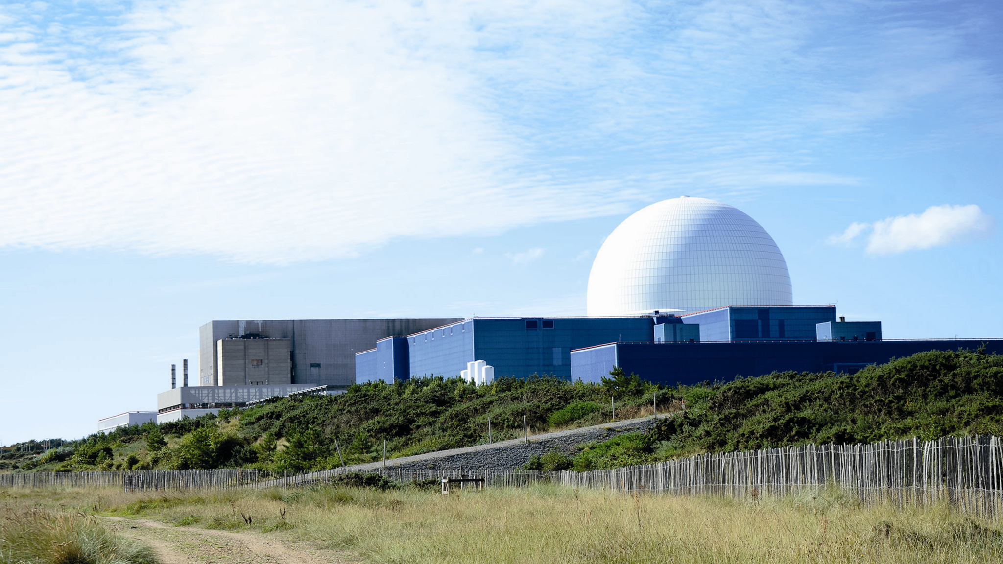 The Sizewell B Power Station on a sunny day. There are large buildings including a dome shaped building. In front of the site are green areas and some footpaths.