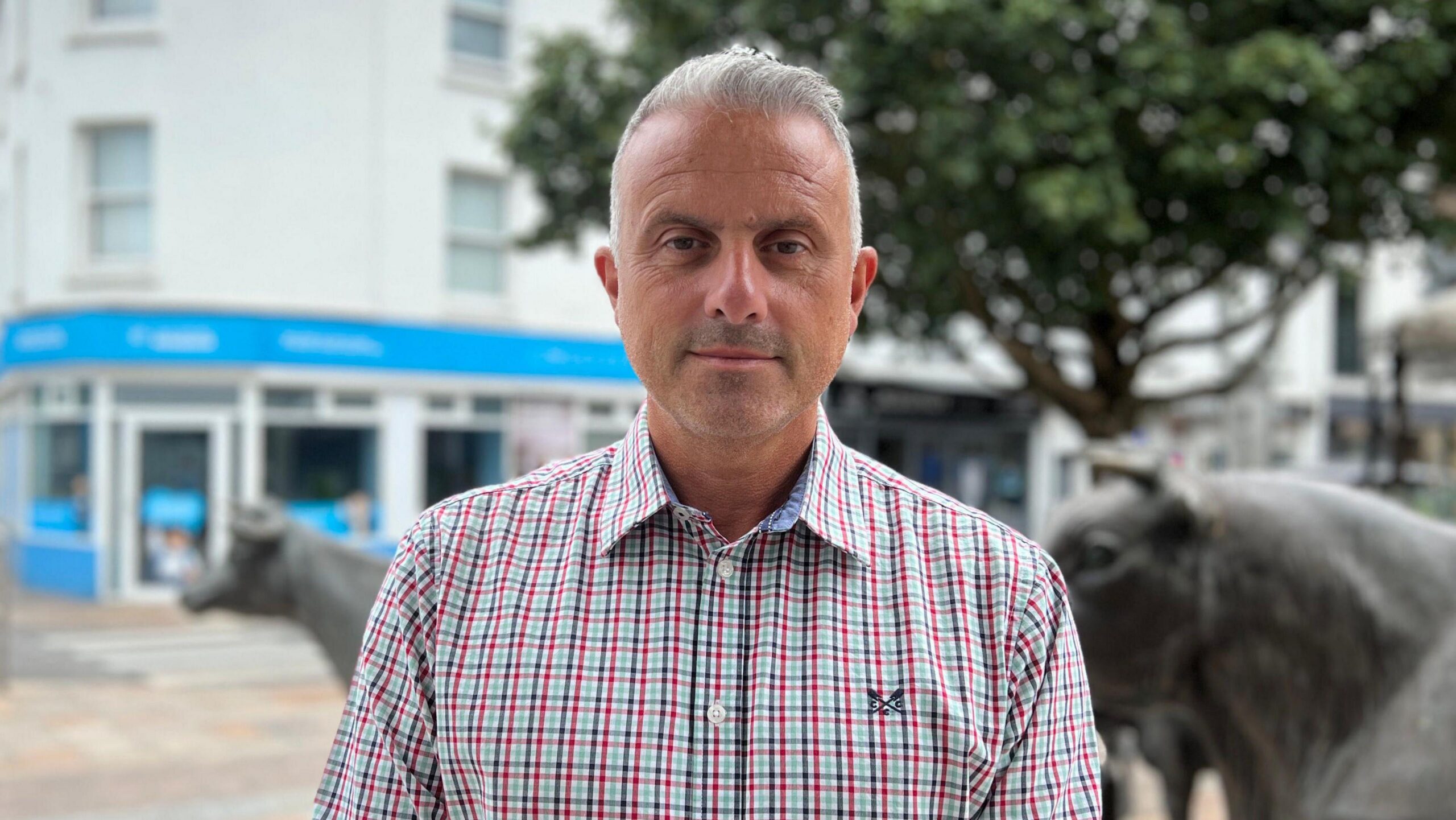 Carl is in a checked button-down shirt stands in a town square, with bronze cattle sculptures, a large tree and shopfronts visible in the blurred background.