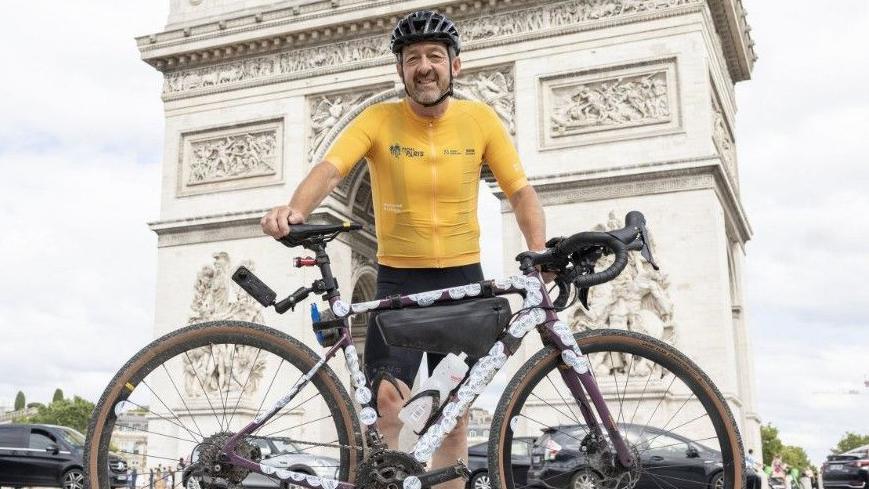 Chris Boardman, who is wearing cycling gear including a helmet, is standing behind his bike and smiling in front of the Arc de Triomphe in Paris.