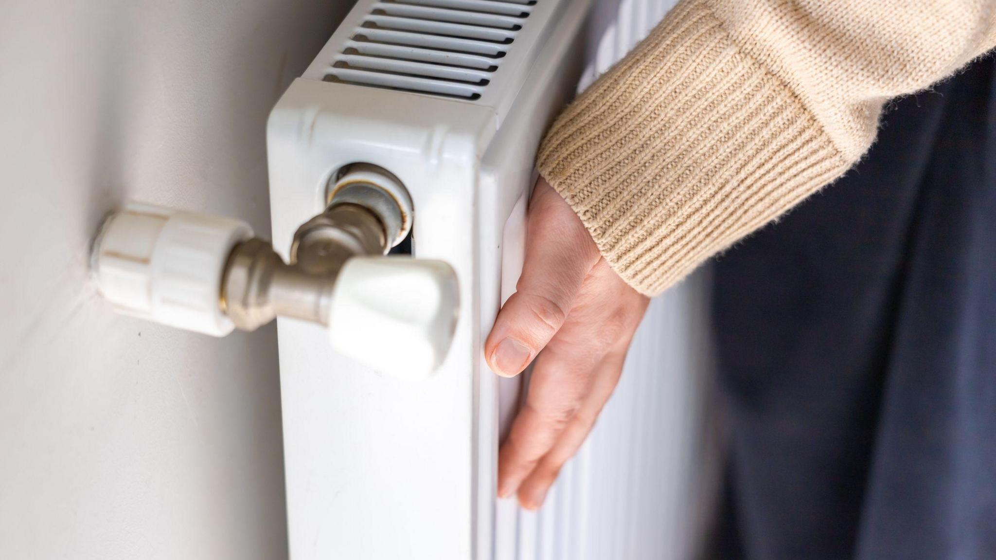 A woman's hand touches a white radiator. The woman's arm and legs can only be seen. She is wearing a cream cardigan and dark trousers.
