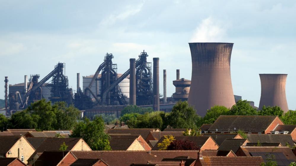 Brown roofs and leafy green trees sit in front of the squat brown blast furnace smokestacks and industrial pipes and silos of the British Steel plant in Scunthorpe.